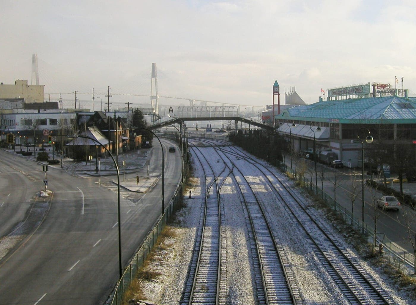 Elevated view of snow-dusted railway tracks between a quiet road and a green-roofed waterfront market, with a pedestrian overpass and cable-stayed bridge towers in the haze