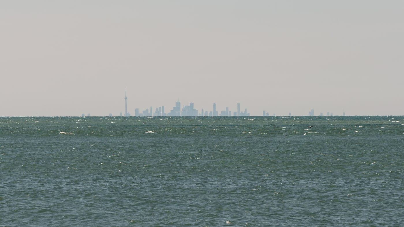 Distant Toronto skyline with the CN Tower on the horizon, seen across choppy green lake water under a hazy pale sky