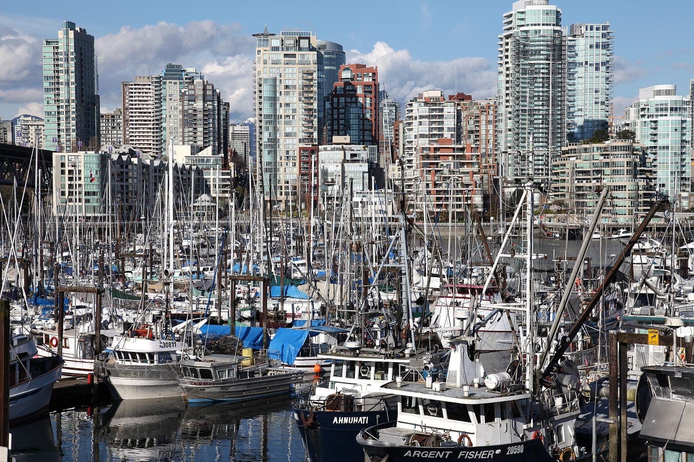 Fishing boats and sailboat masts crowd a marina in the foreground, with tall glass and brick condo towers rising behind under a blue sky with scattered clouds.