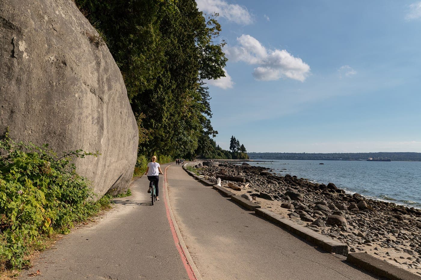 A cyclist rides along a paved seaside path beside a massive rock face and tall evergreen trees, with a rocky beach, calm ocean and a distant freighter under a blue sky.