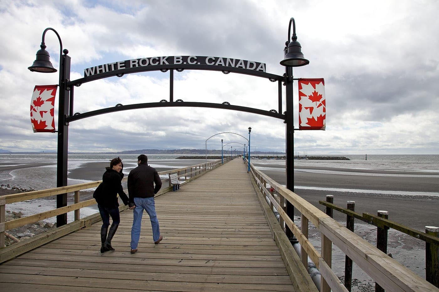 A couple walks along a long wooden pier under an arch reading White Rock B.C. Canada, with maple leaf banners, low-tide sand flats and cloudy sky